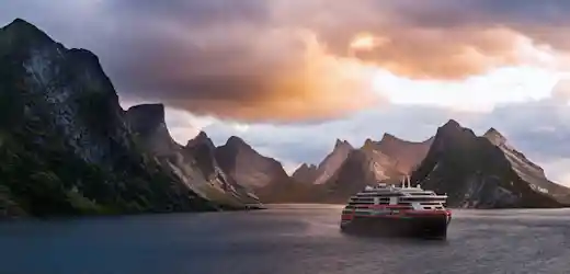 Cruise ship sailing in fjord surrounded by jagged mountains under orange sunset clouds