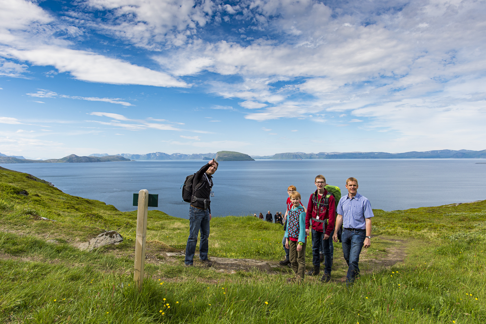 Hikers on a grassy coastal bluff overlooking a fjord with distant islands and blue sky