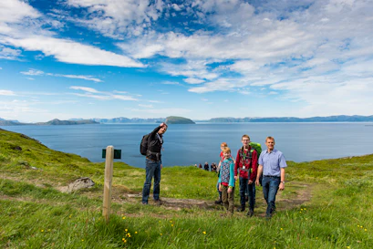 Hikers on a grassy coastal bluff overlooking a fjord with distant islands and blue sky