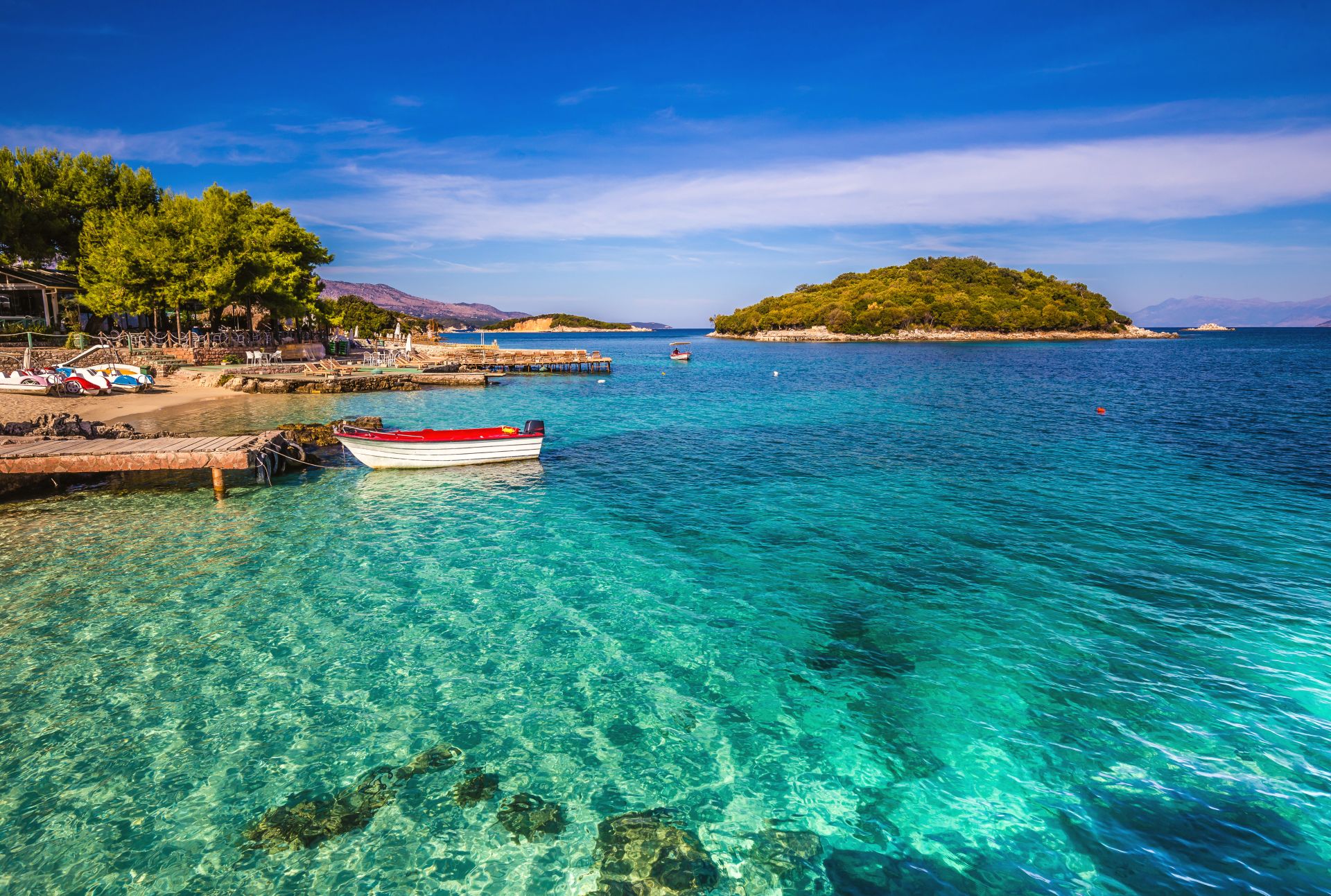 Small red-and-white boat by a wooden pier, turquoise bay with a tree-lined shore and island.