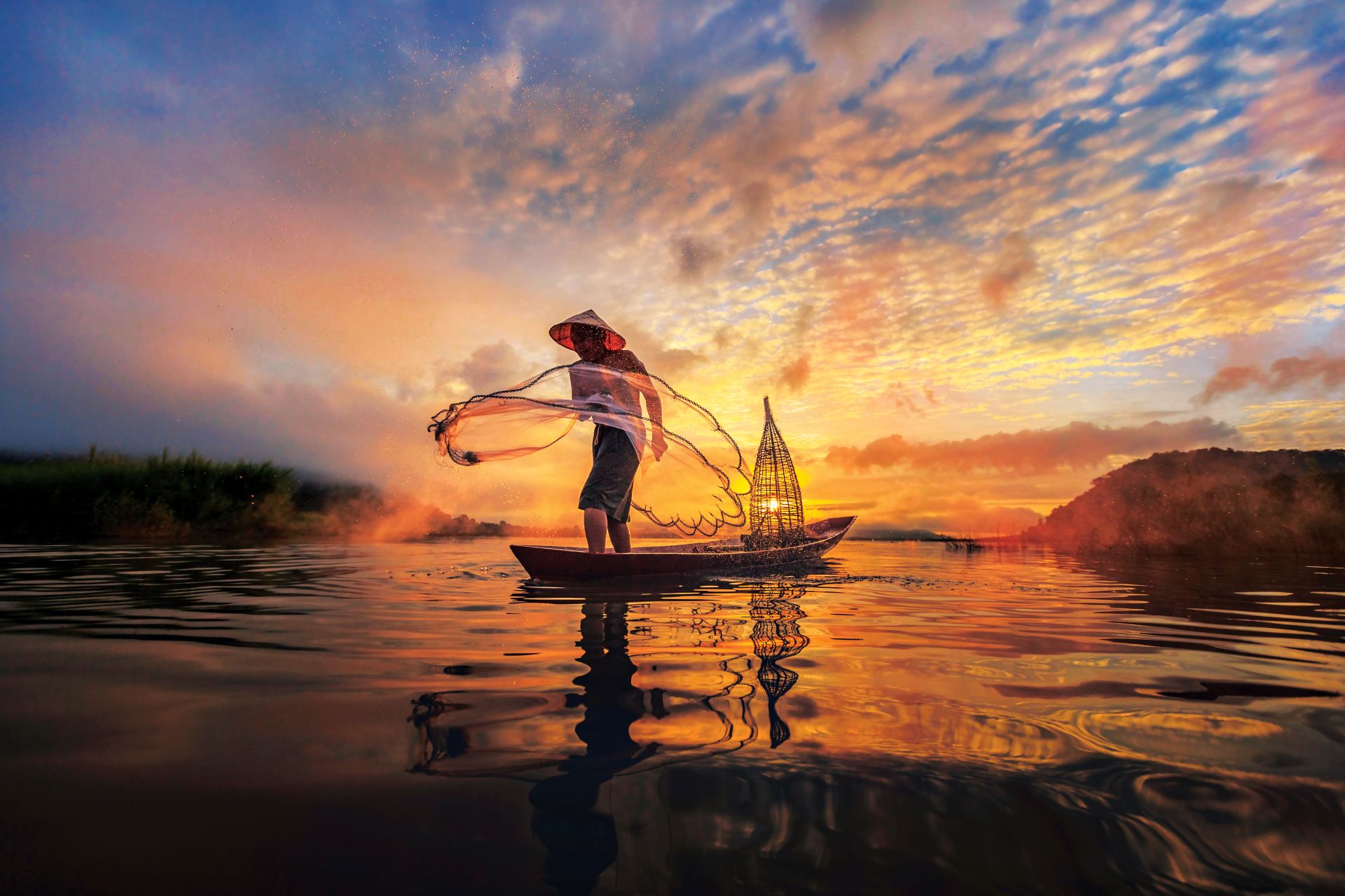 Fisherman casting a circular net from a small boat at golden sunset with woven trap and reflections