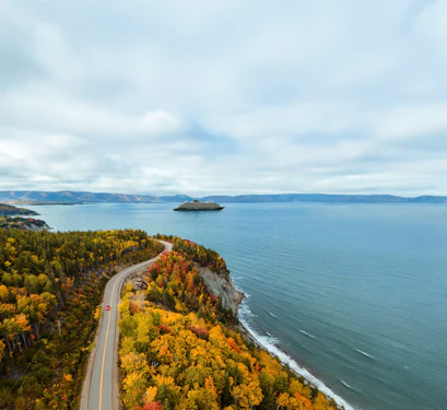 Aerial view of autumn coastline with winding road and red car, cruise ship on calm bay