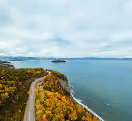 Aerial view of autumn coastline with winding road and red car, cruise ship on calm bay