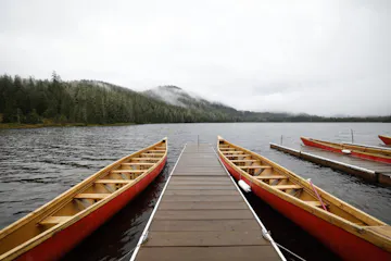 Wooden dock between red canoes on a calm lake with forested shore and distant mountains in mist