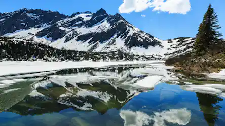 Snow-covered peaks reflected in a partially frozen alpine lake with evergreen tree
