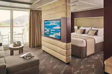 Cruise-suite living area with sofa and breakfast table, partitioned TV, king bed and balcony view.