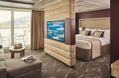 Cruise-suite living area with sofa and breakfast table, partitioned TV, king bed and balcony view.