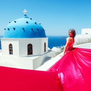 Woman in flowing red gown on white terrace beside blue-domed church overlooking sea.
