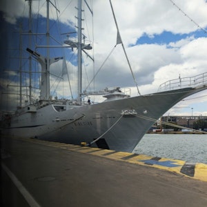 Large white sailing ship moored at harbor, bow in foreground, tall masts under cloudy sky