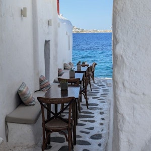 Greek narrow seaside alley café with wooden tables, cushions, and stone path to blue sea