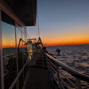 Sunset over open sea from cruise deck, railing and lifeboat reflected in windows
