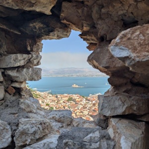 View through stone wall opening onto coastal town of red roofs, blue sea, small island fortress.