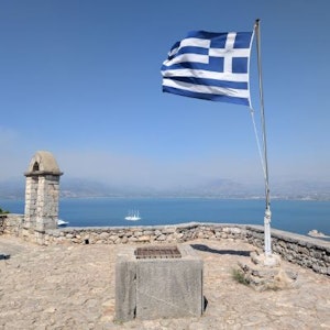 Greek flag on a hilltop stone fort overlooking blue sea, distant coast and sailing ship