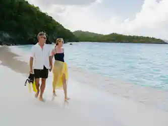 Smiling couple in sunglasses holding hands, walking barefoot on tropical beach with snorkeling gear
