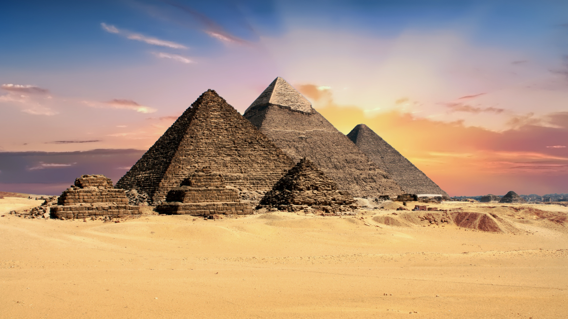 Three Great Pyramids of Giza with smaller mastabas on a sandy plain beneath a colorful sunset sky