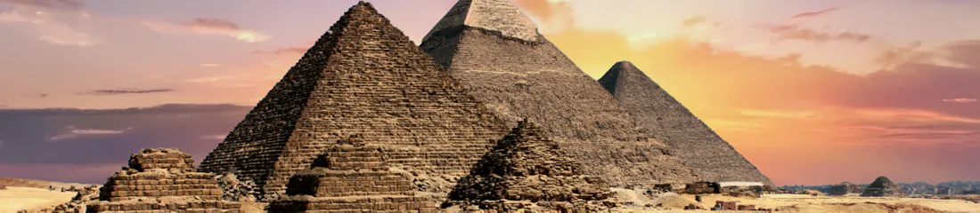 Three Great Pyramids of Giza with smaller mastabas on a sandy plain beneath a colorful sunset sky