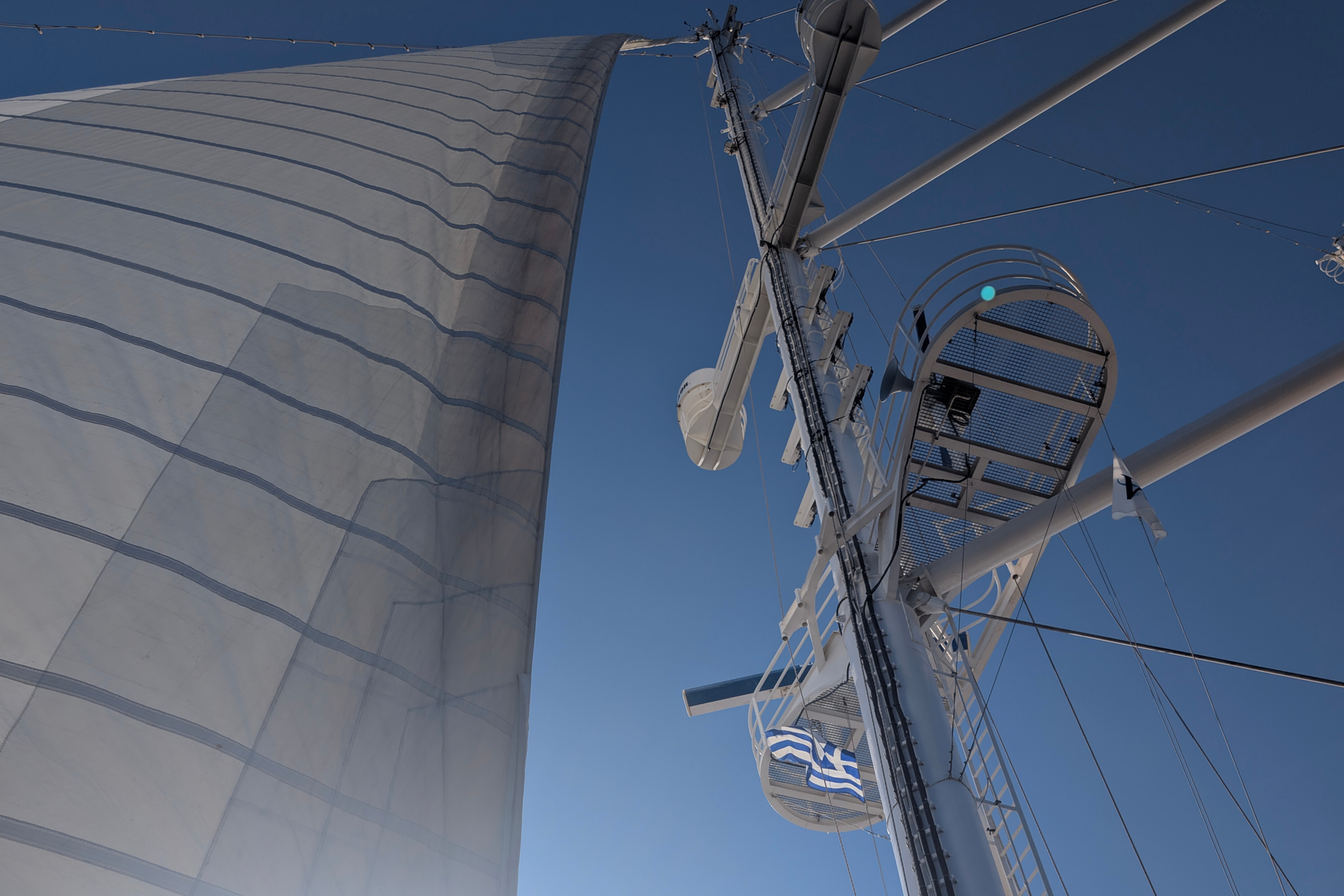 Upward view of yacht mast, rigging, large seamed sail and Greek flag