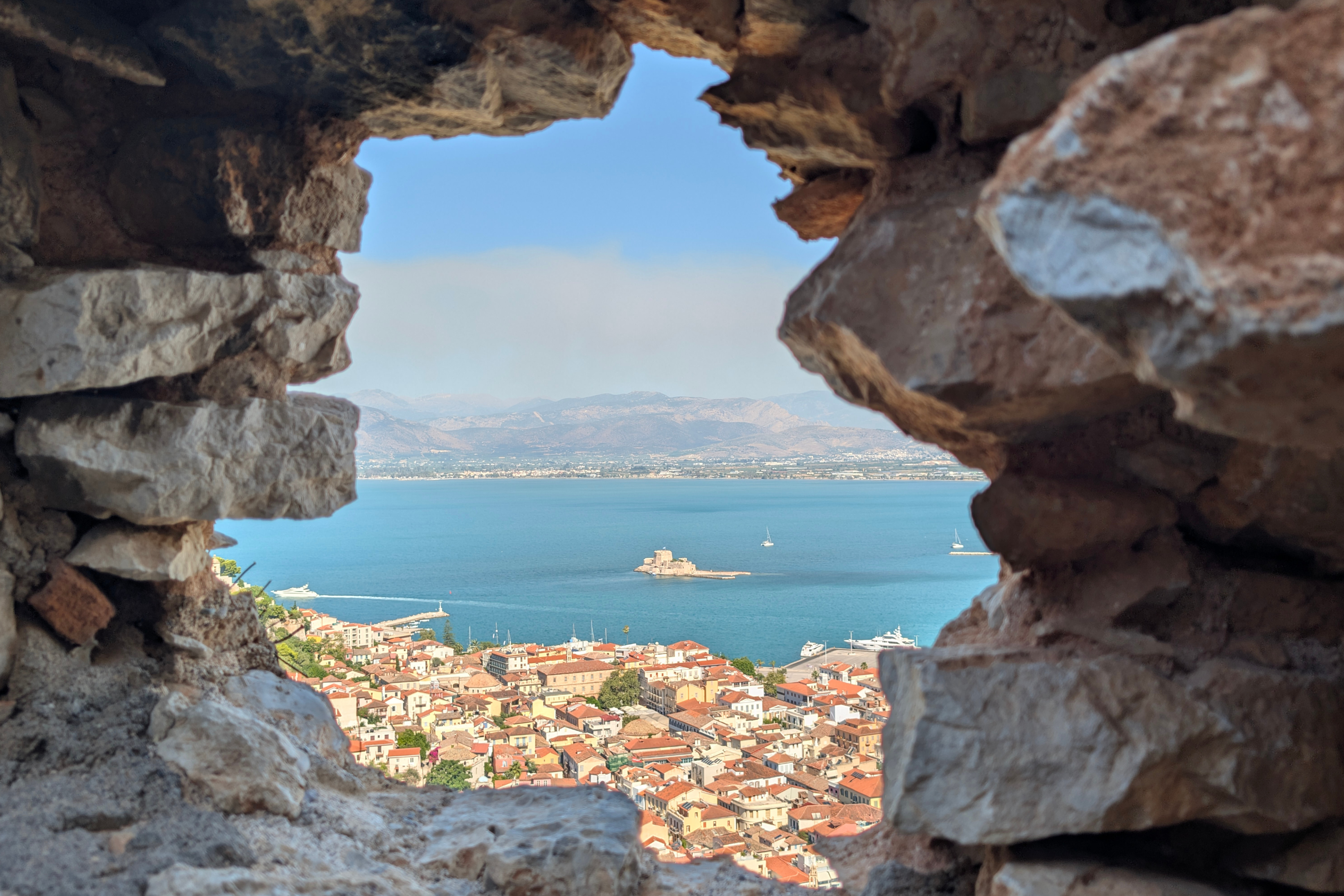 Stone arch framing red-roofed coastal town, harbor, tiny island fortress and distant mountains