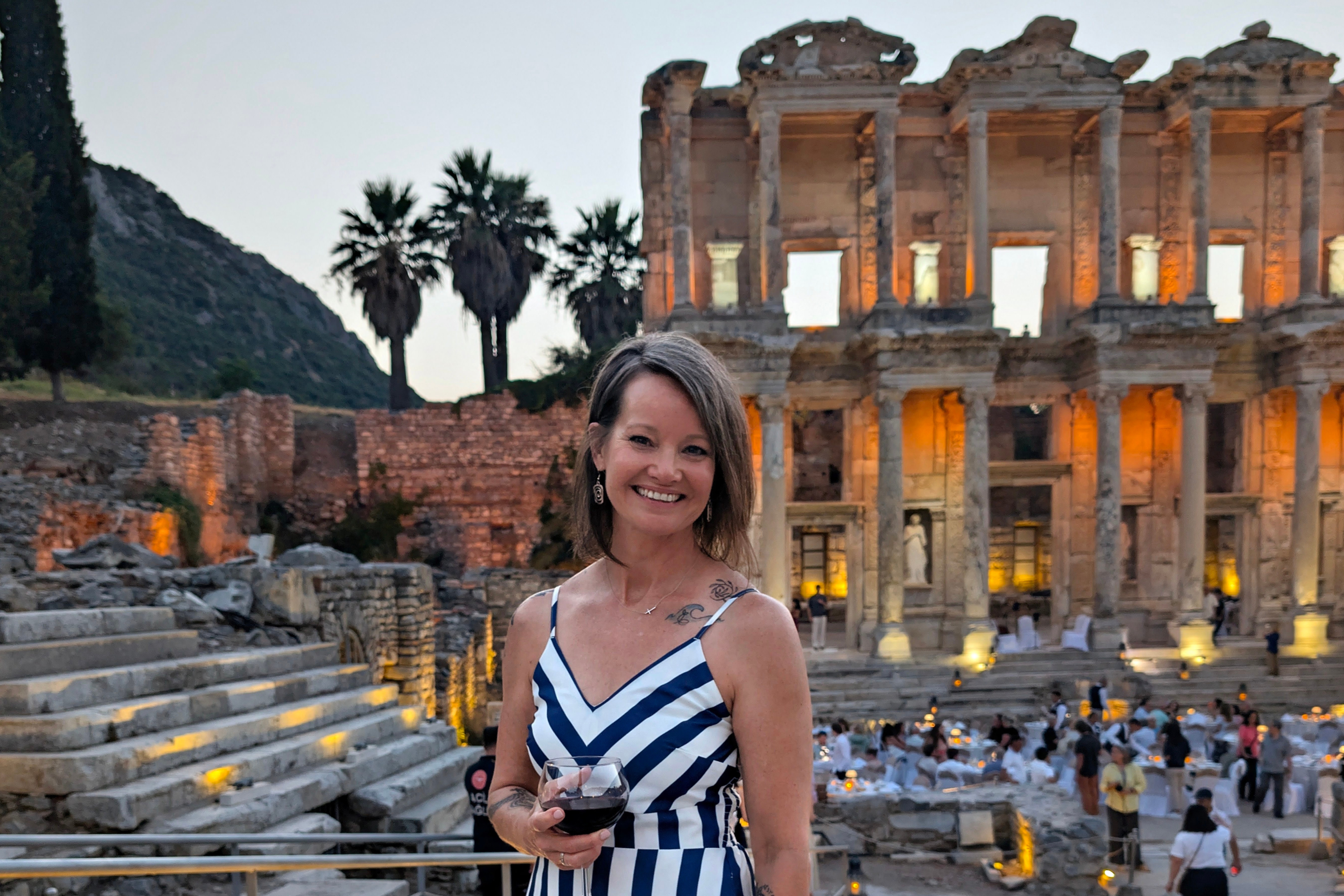 Smiling woman in blue-white striped dress holding wine before illuminated ancient ruins