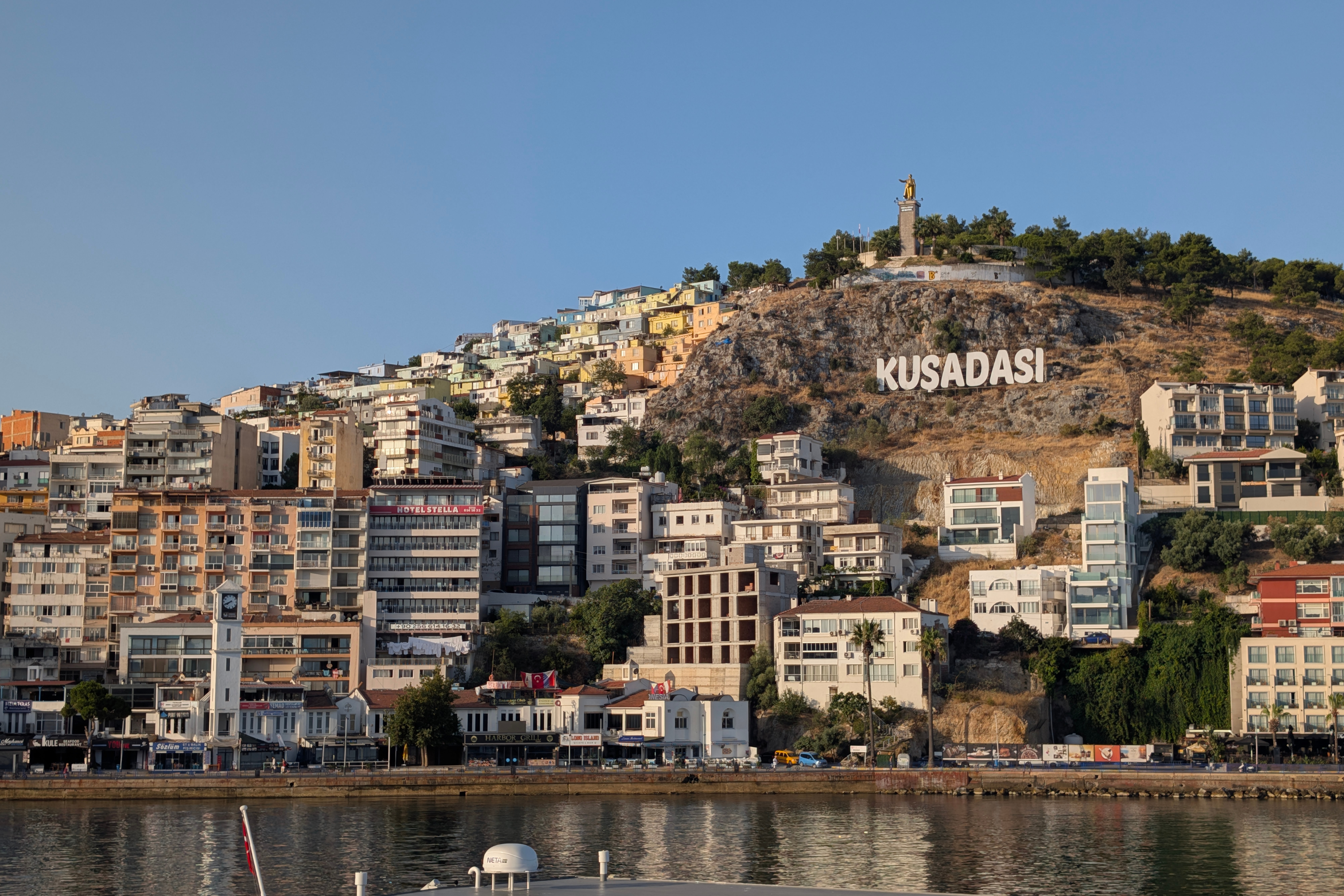 Kuşadası hillside with white sign, stacked pastel houses, waterfront and hilltop statue