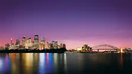 Sydney skyline at dusk: lit skyscrapers, Sydney Opera House and Harbour Bridge reflected in harbor