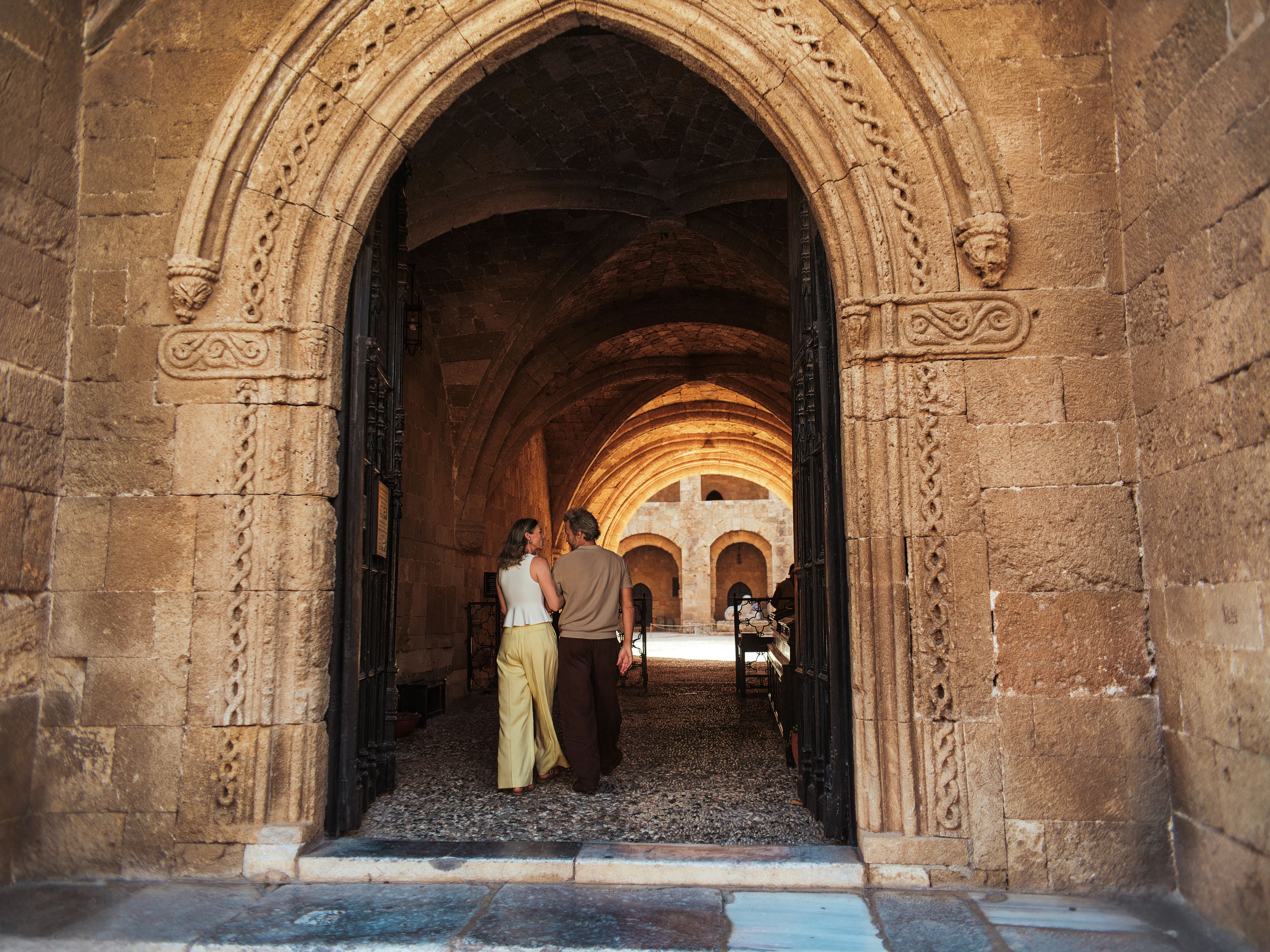 Couple walking arm-in-arm through carved stone arch into sunlit vaulted cloister