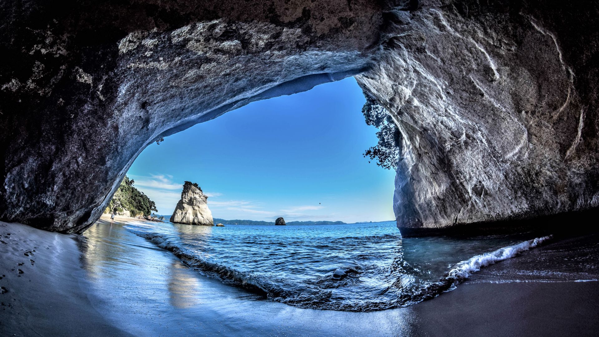 Inside sea cave framing sandy shore, gentle waves and a solitary rock stack under blue sky