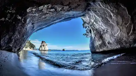 Inside sea cave framing sandy shore, gentle waves and a solitary rock stack under blue sky