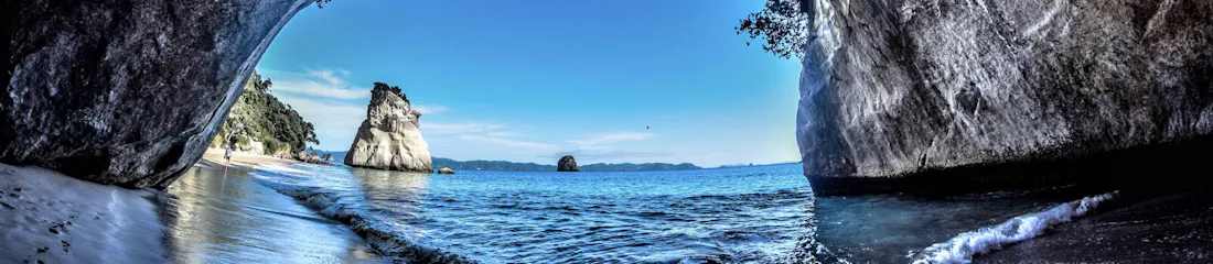 Inside sea cave framing sandy shore, gentle waves and a solitary rock stack under blue sky