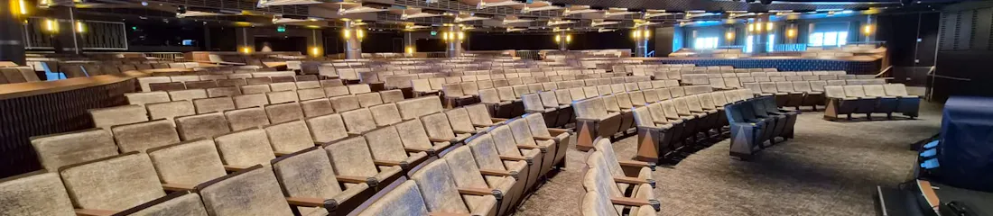 Empty curved auditorium with rows of upholstered seats, overhead lights and stage rigging