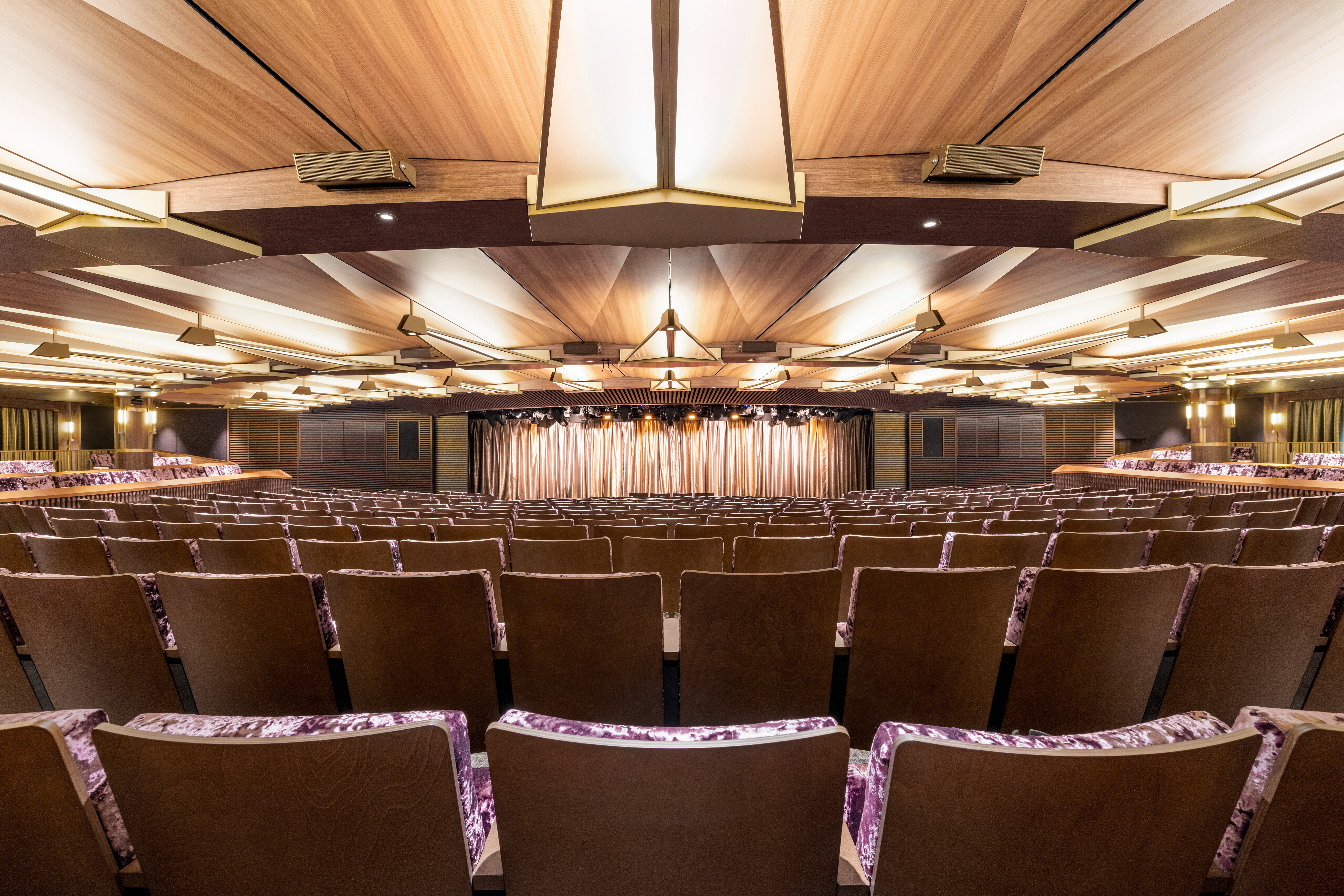 Empty theater auditorium with rows of seats, wooden geometric ceiling, and stage curtain