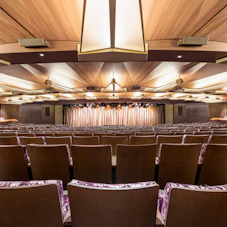 Empty theater auditorium with rows of seats, wooden geometric ceiling, and stage curtain