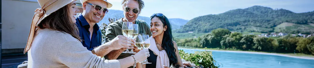 Four smiling friends toasting white wine on a stone terrace overlooking a river and green hills