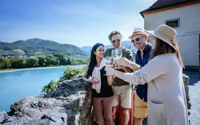Four friends toasting white wine on a stone terrace overlooking a river and green hills