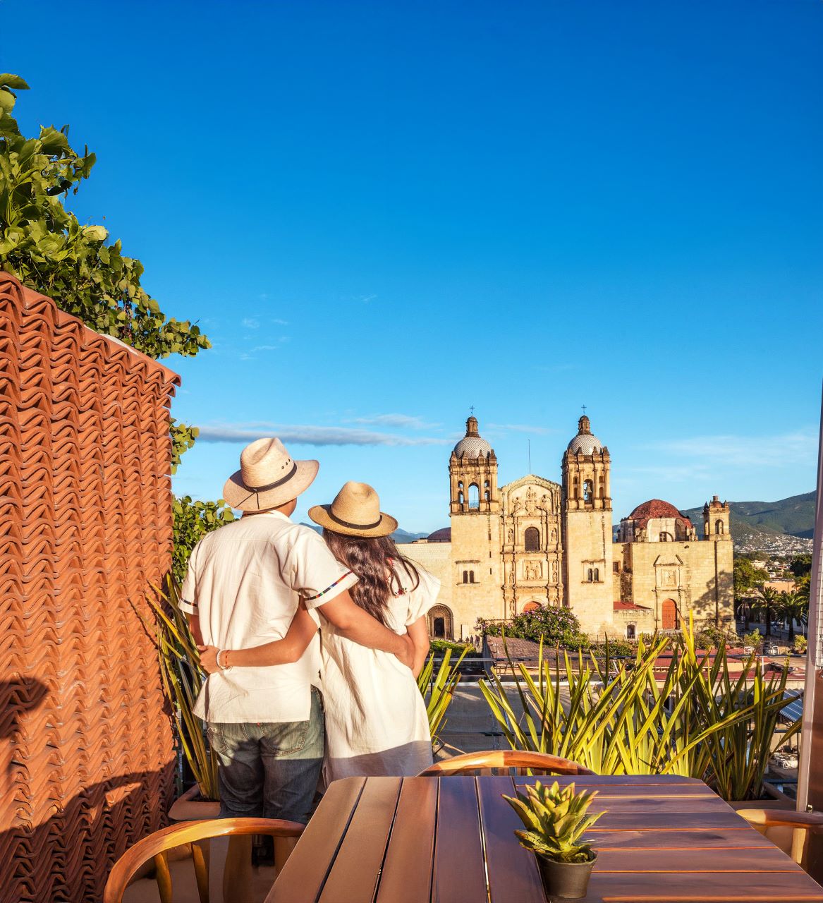 Couple in straw hats embracing on rooftop terrace overlooking ornate cathedral and blue sky