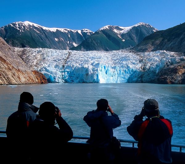 Four people on a boat using binoculars to view a blue glacier and snow-capped mountains