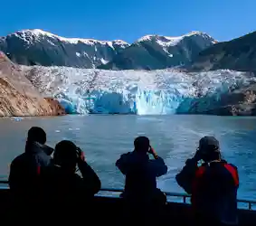 Four people on a boat using binoculars to view a blue glacier and snow-capped mountains