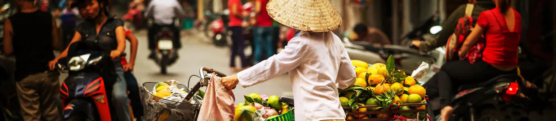 Vendor with conical hat pushes bicycle laden with oranges amid busy motorbikes on market street