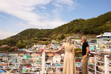 Man and woman on rooftop railing overlooking colorful hillside village and forested hill