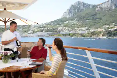 Waiter pouring red wine for a couple dining on a yacht deck with hillside coastal town