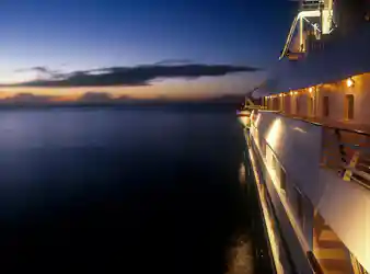 Lit cruise ship starboard deck with lifeboat at dusk, calm sea and orange-blue horizon