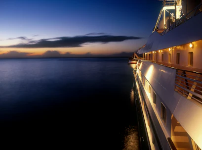 Lit cruise ship starboard deck with lifeboat at dusk, calm sea and orange-blue horizon