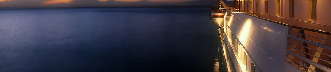 Lit cruise ship starboard deck with lifeboat at dusk, calm sea and orange-blue horizon