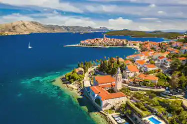 Walled Mediterranean town on a red-roofed peninsula, turquoise sea, sailboat and distant islands