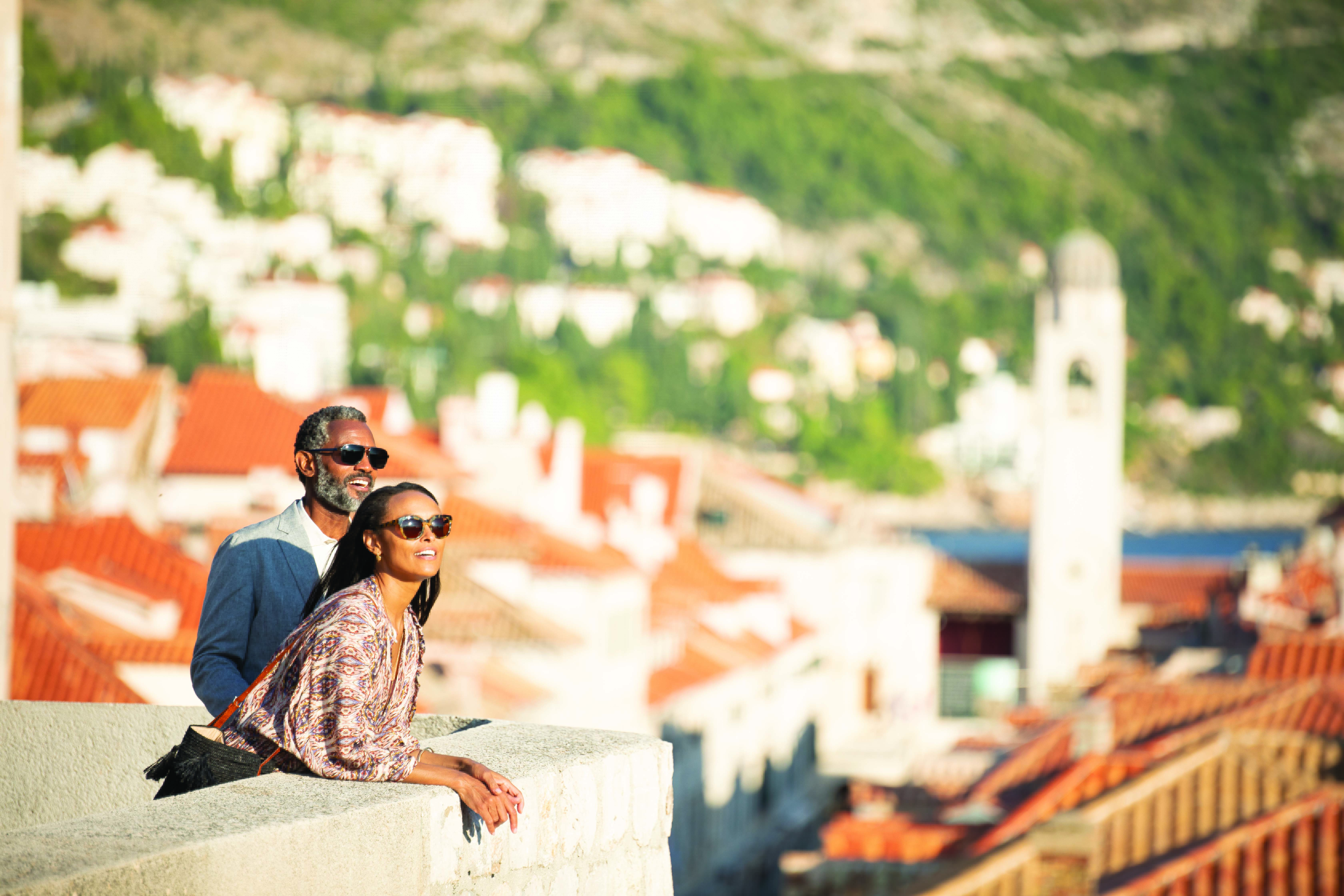 Couple in sunglasses leaning on a stone wall overlooking red-tiled coastal town and bell tower