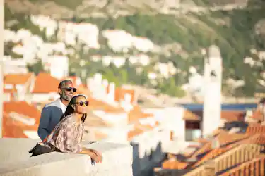 Couple in sunglasses leaning on a stone wall overlooking red-tiled coastal town and bell tower