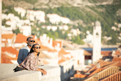 Couple in sunglasses leaning on a stone wall overlooking red-tiled coastal town and bell tower