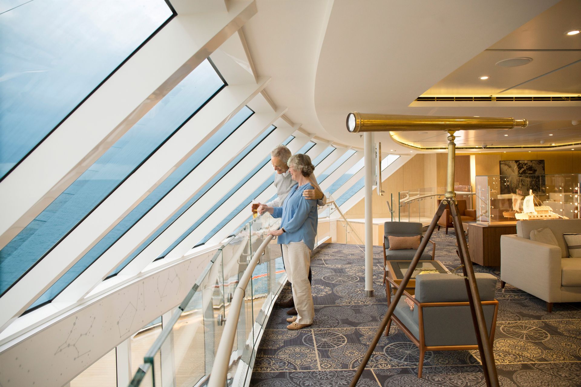 Elderly couple at glass railing looking out ocean through slanted cruise-ship windows in lounge