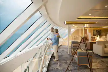 Elderly couple at glass railing looking out ocean through slanted cruise-ship windows in lounge