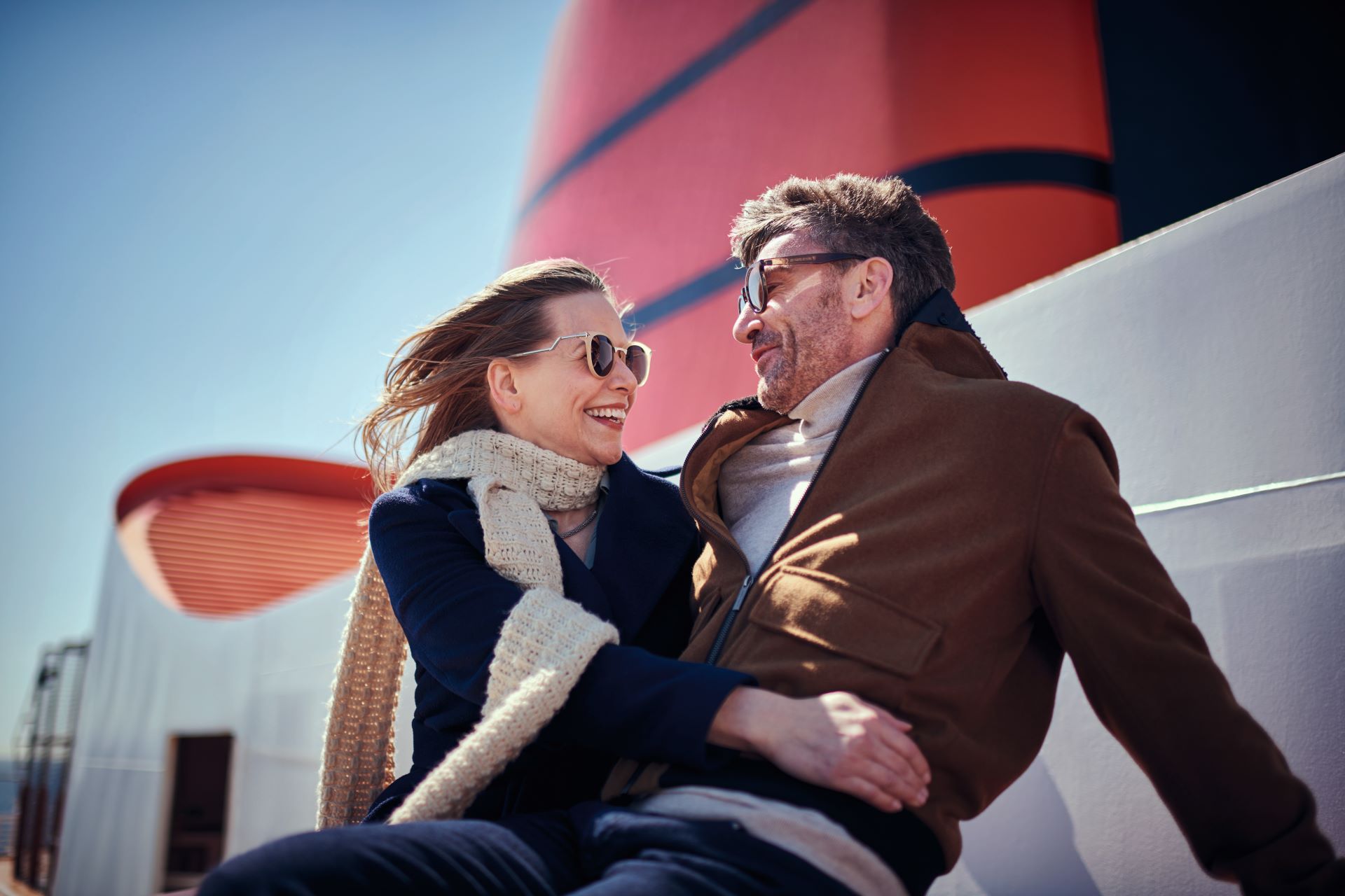 Laughing couple in sunglasses embrace on windy ship deck beside a tall red smokestack